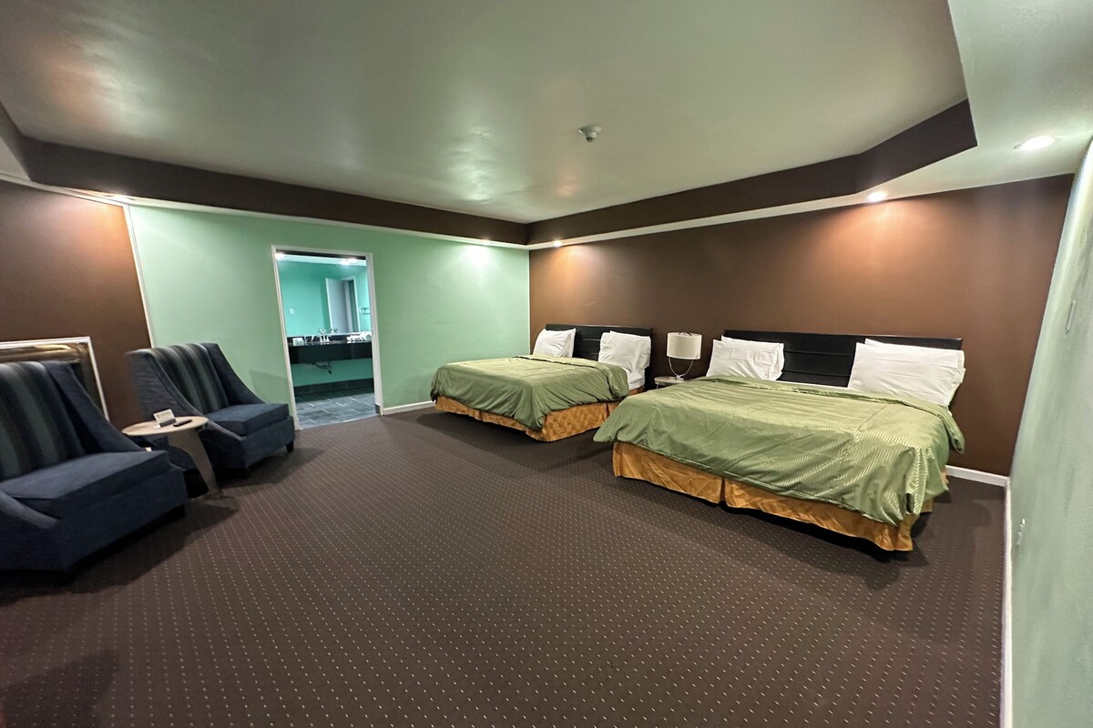Habitación de hotel con dos camas individuales, un sofá, una mesita y un baño visible a través de una puerta abierta.