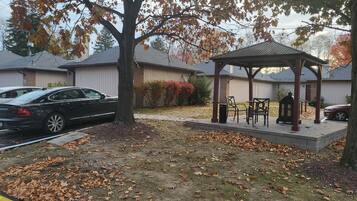 A gazebo with chairs and a table, surrounded by a grassy area with fallen leaves.