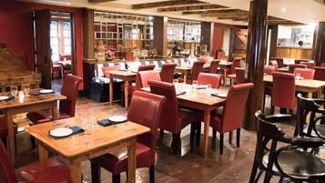 A restaurant with wooden tables and red chairs, featuring a brick wall and exposed wooden beams on the ceiling.