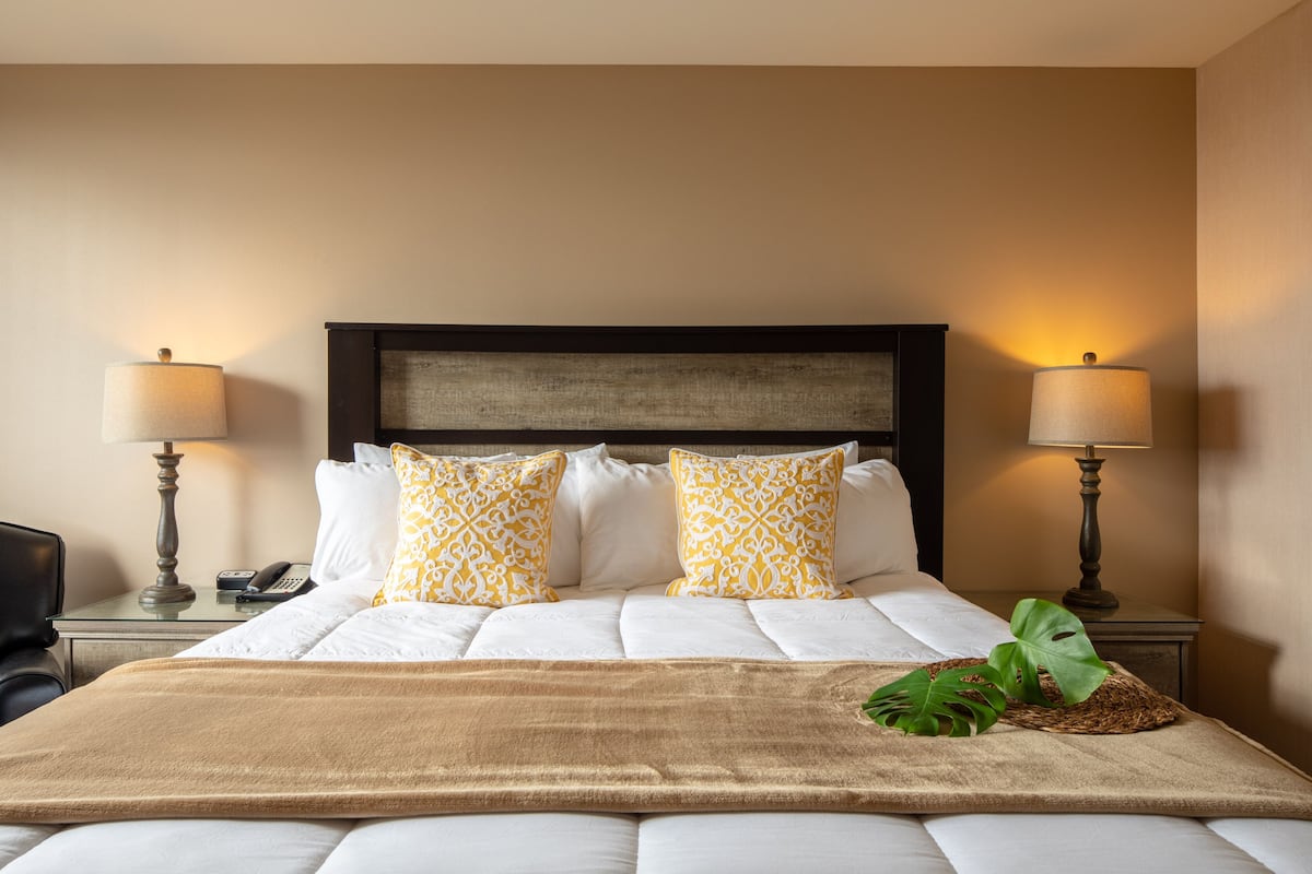 A neatly made bed with white and yellow patterned pillows, a brown throw blanket, and a plant on the bed. There are two bedside lamps on either side of the bed.