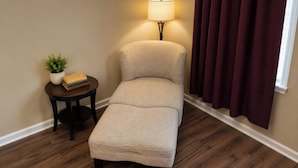 A small, single-tiered sofa with a light-colored fabric, a round wooden side table with a plant and books, a floor lamp, and a window with dark curtains. - Plantation Oaks Suites & Inn Millington (Millington)