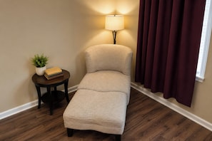 A small, single-tiered sofa with a light-colored fabric, a round wooden side table with a plant and books, a floor lamp, and a window with dark curtains. - Plantation Oaks Suites & Inn Millington (Millington)