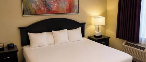 A hotel room with a bed, a nightstand with a lamp, a painting on the wall, and a window with curtains.