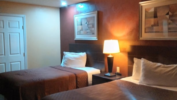 Habitación de hotel con dos camas, una mesita de noche con lámpara, un cuadro en la pared y una puerta.
