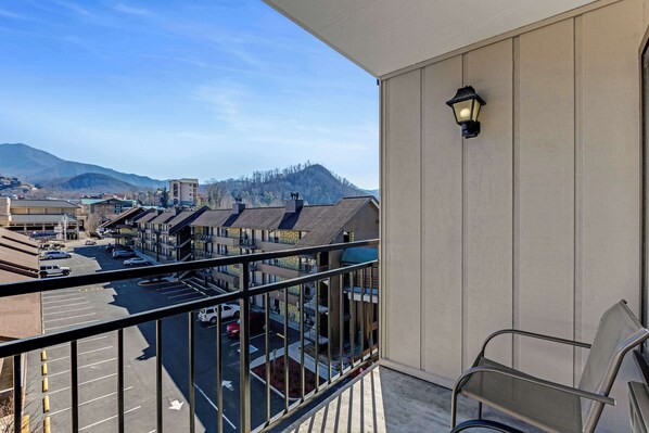 A balcony with a view of a parking lot and buildings, a chair, and a wall-mounted light fixture.