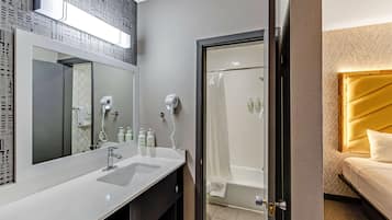 A modern bathroom with a large mirror, dual sinks, a shower, and a bed with a headboard.