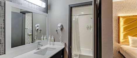 A modern bathroom with a large mirror, dual sinks, a shower, and a bed with a headboard.