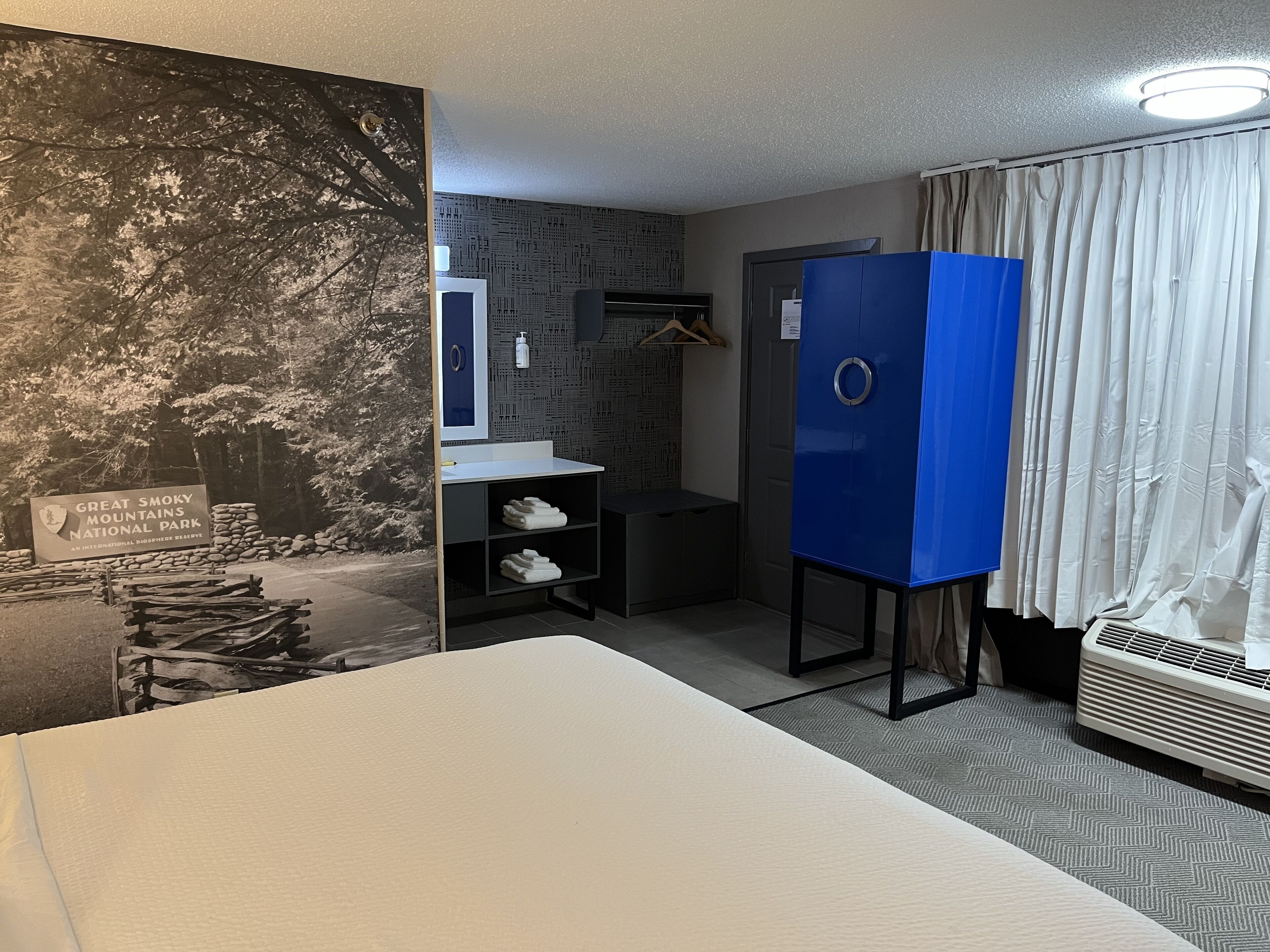 A hotel room with a bed, a bathroom with a sink and mirror, a blue wardrobe, and a poster of the Great Smoky Mountains National Park.