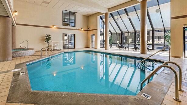 An indoor swimming pool with clear glass walls, a tiled floor, and a sunlit patio area with outdoor seating.
