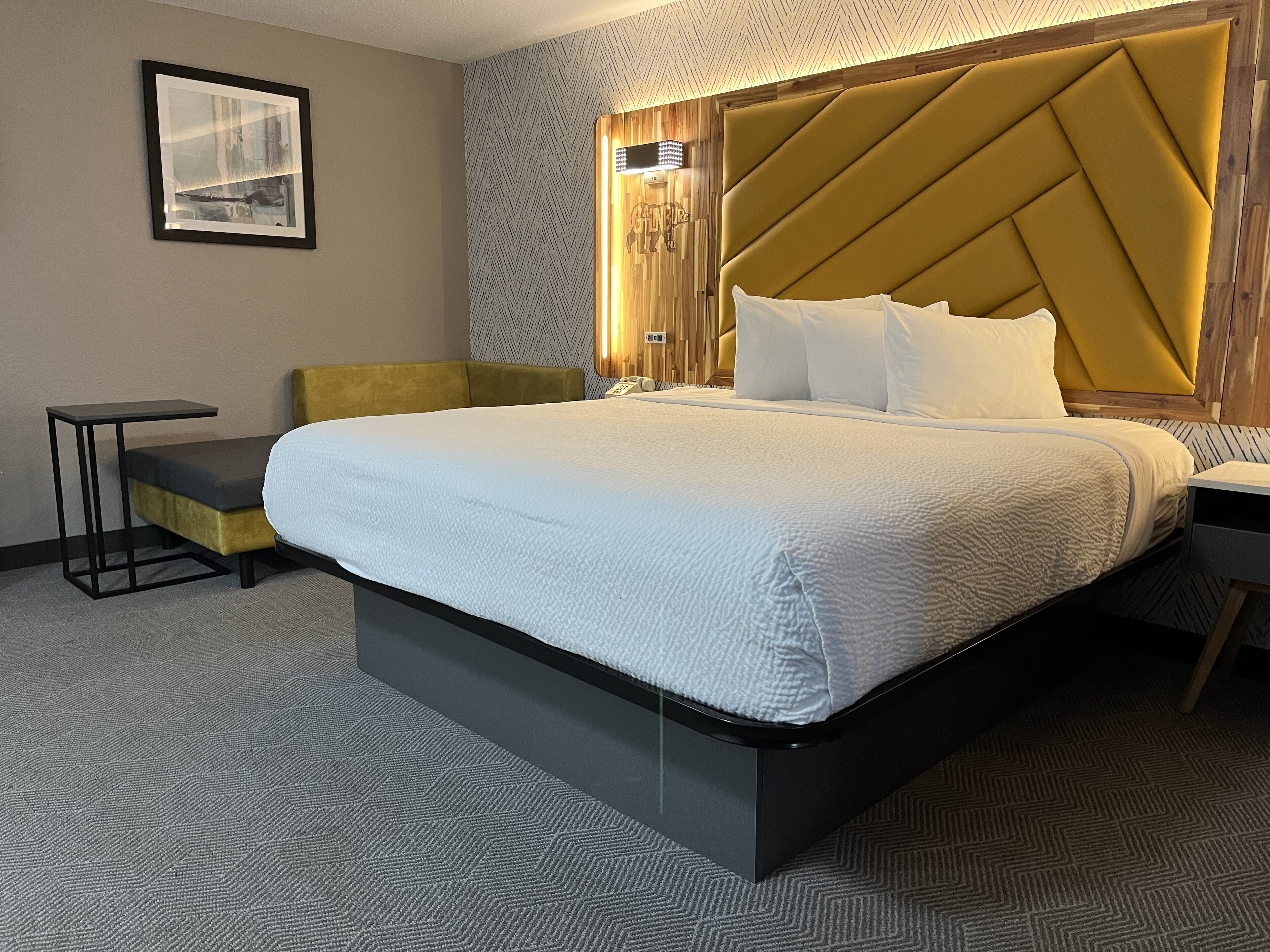 A hotel room with a large bed, a small side table, a sofa, and a headboard with a light fixture.
