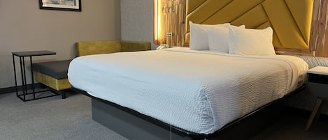 A hotel room with a large bed, a small side table, a sofa, and a headboard with a light fixture.