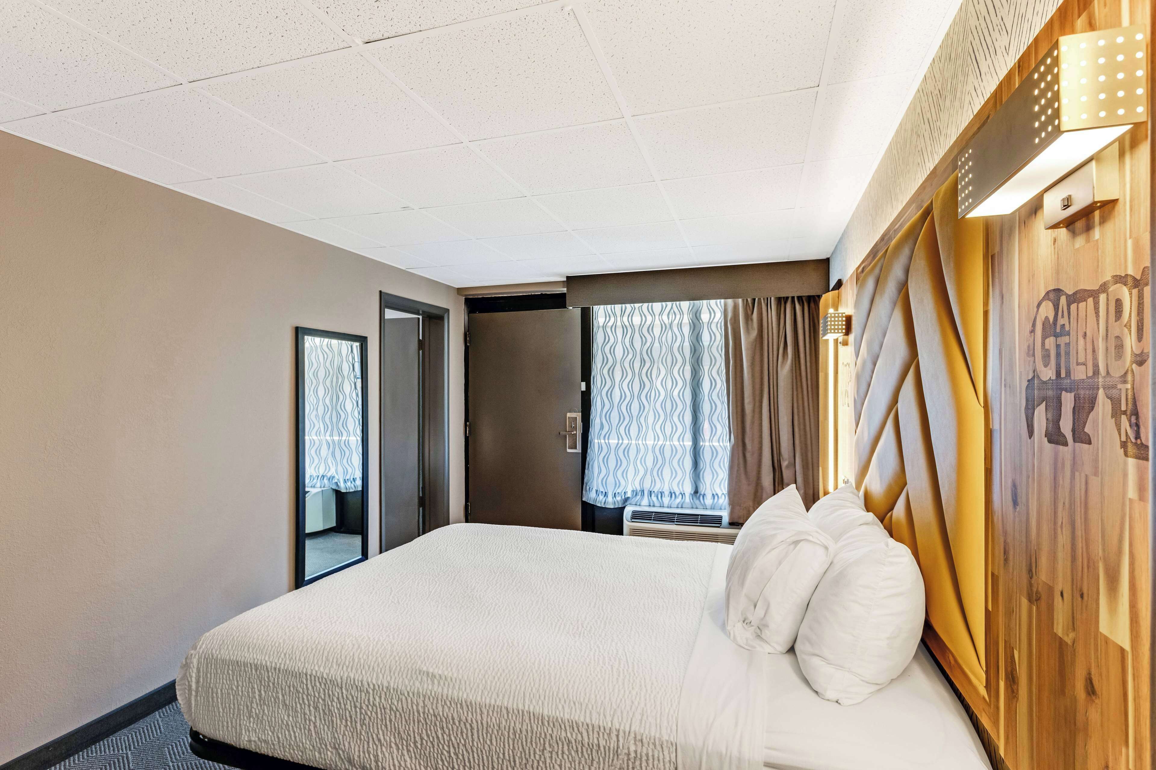 A hotel room with a large bed, a wooden headboard, a window with curtains, and a door leading to another room.
