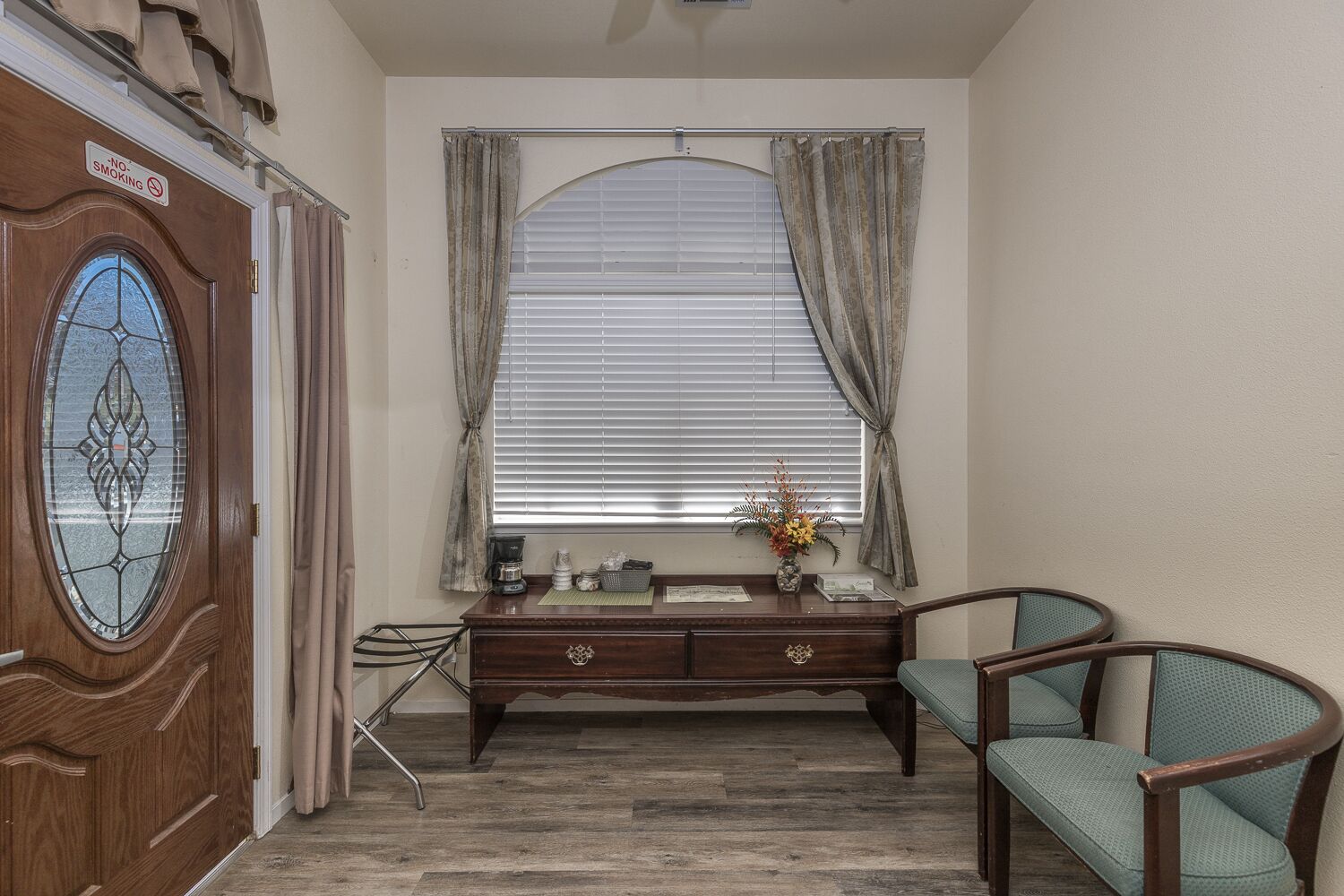 A room with a wooden desk, a chair, a window with curtains, a coffee maker, and a vase with flowers.