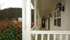 A covered porch with white railings, a wooden floor, and a view of a forested hillside. - 5th Street Inn (Mariposa)