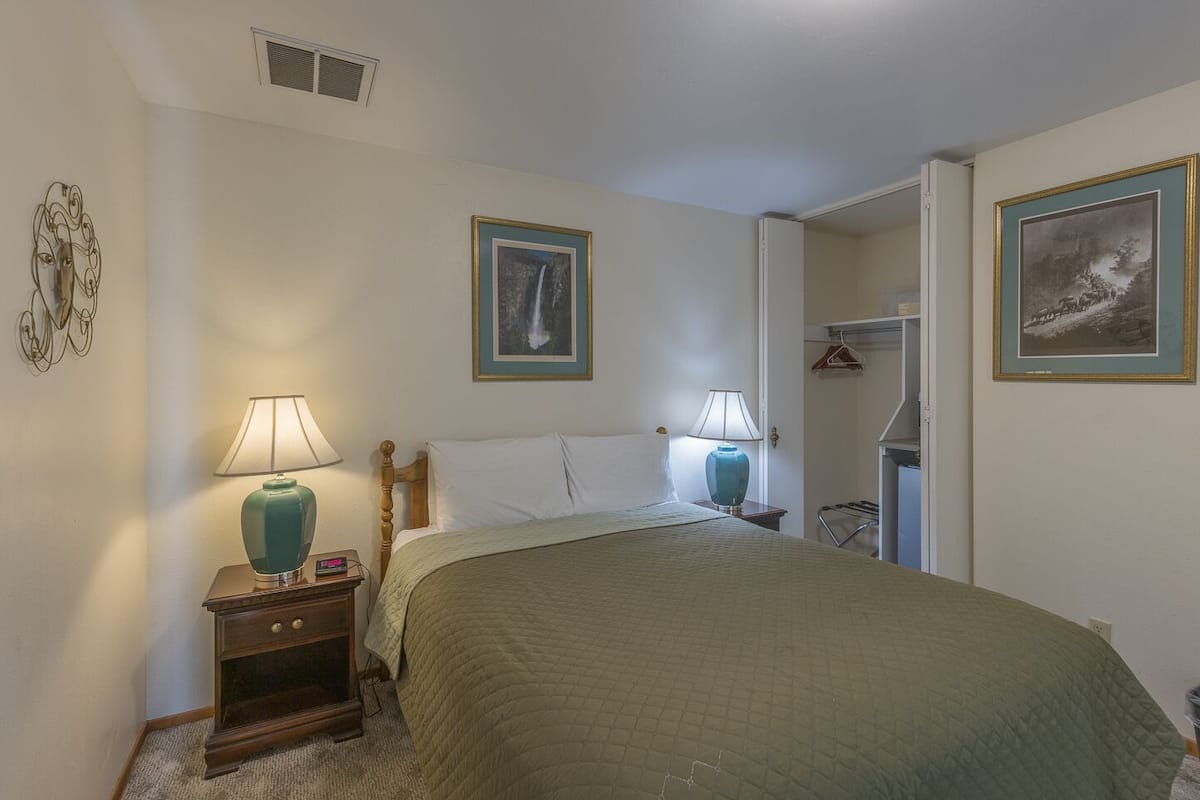 A bedroom with a bed, two bedside lamps, a wooden nightstand, a wall-mounted coat rack, and two framed pictures.