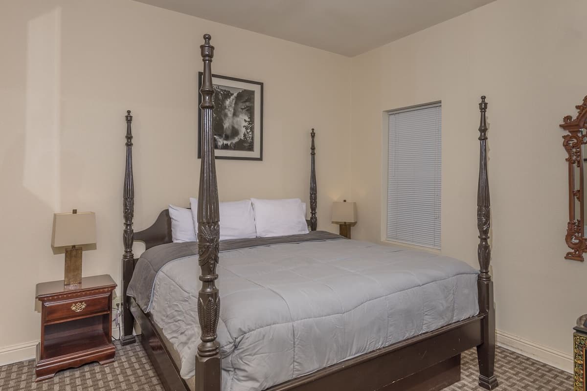 A four-poster bed with a grey bedspread, two bedside tables with lamps, a framed picture on the wall, and a window with blinds.
