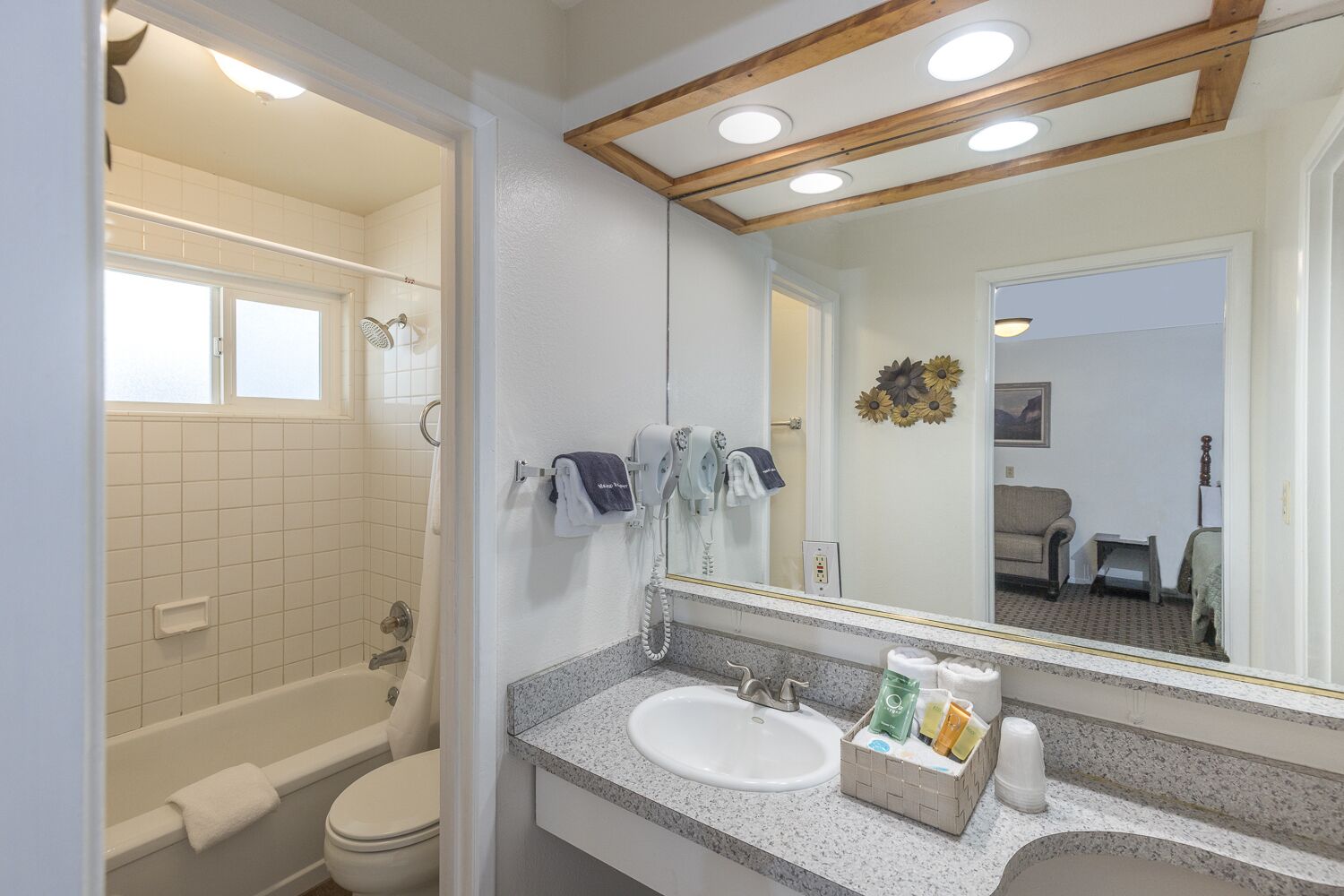 A bathroom with a bathtub, toilet, sink, and a mirror reflecting a living room with a couch and a vase.