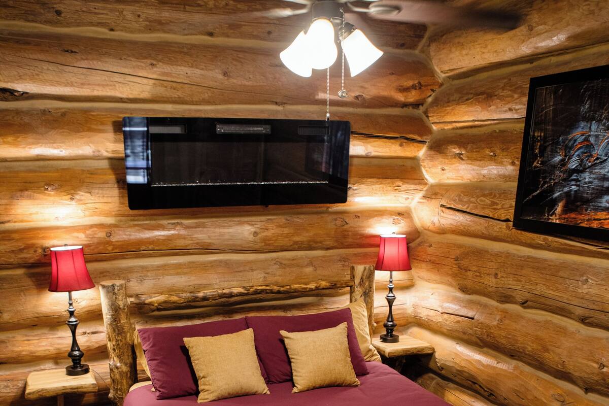 A log cabin room with a wooden bed, two bedside lamps, a ceiling fan, and a mounted television.