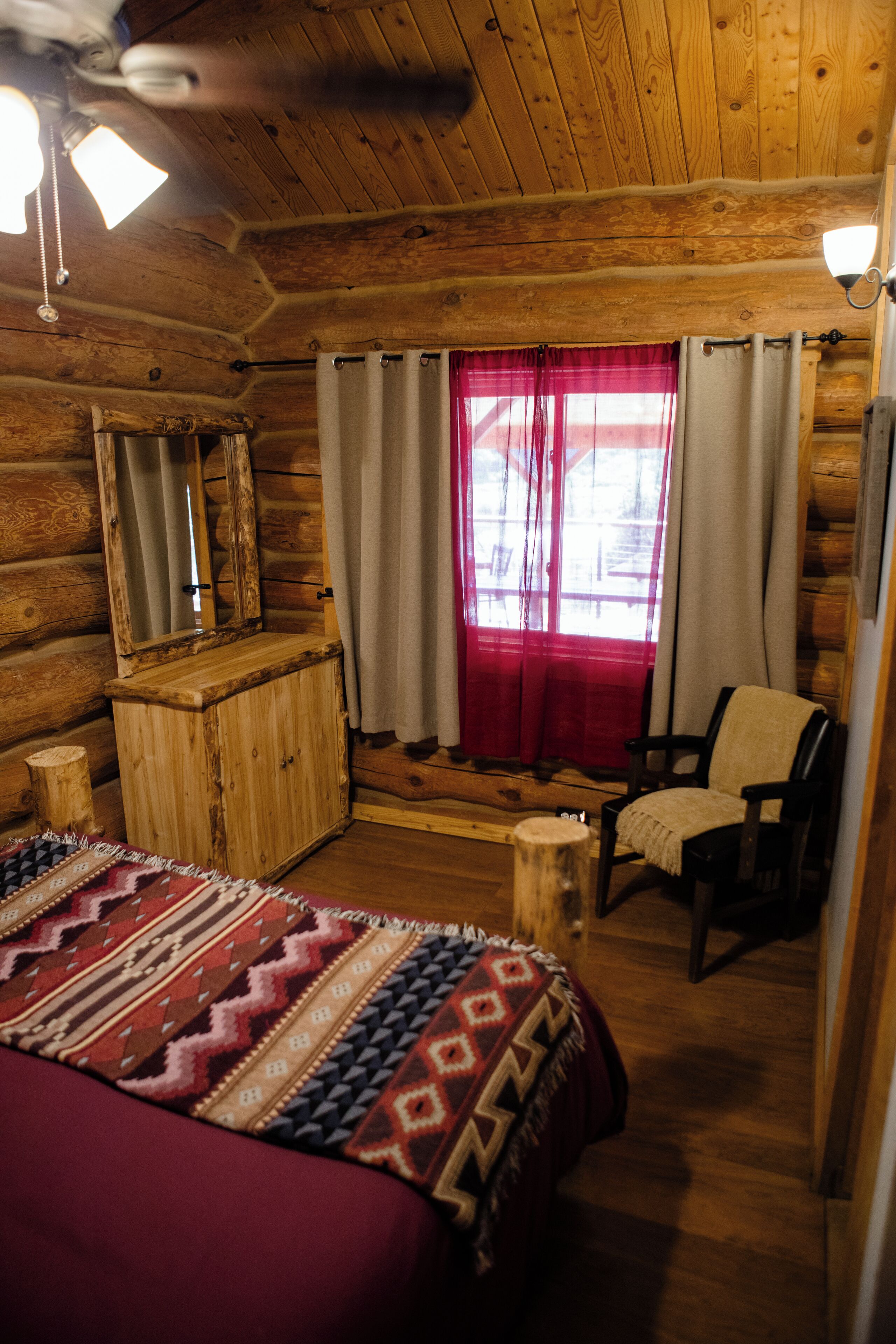 A log cabin room with a bed, a chair, a wooden dresser, and a window with curtains.
