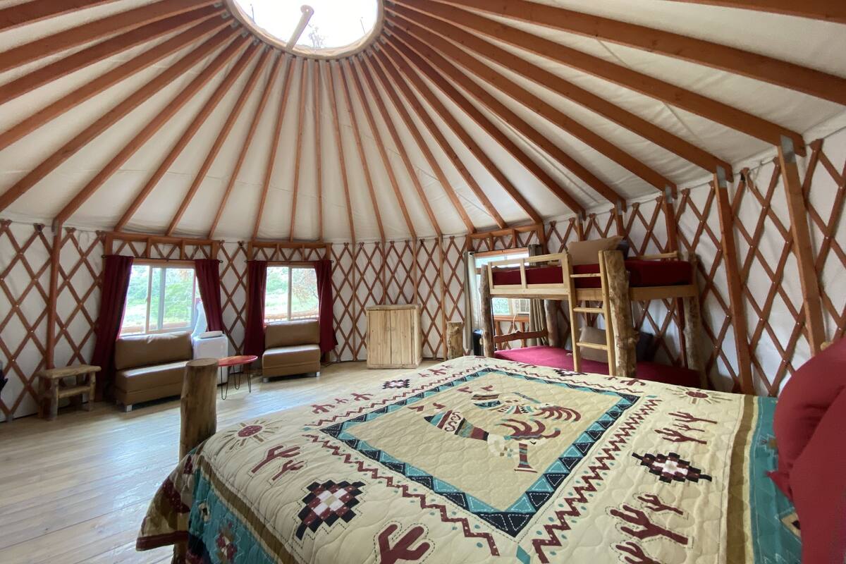 A yurt with a wooden interior, a central bed with a patterned blanket, a small table, and a bench.