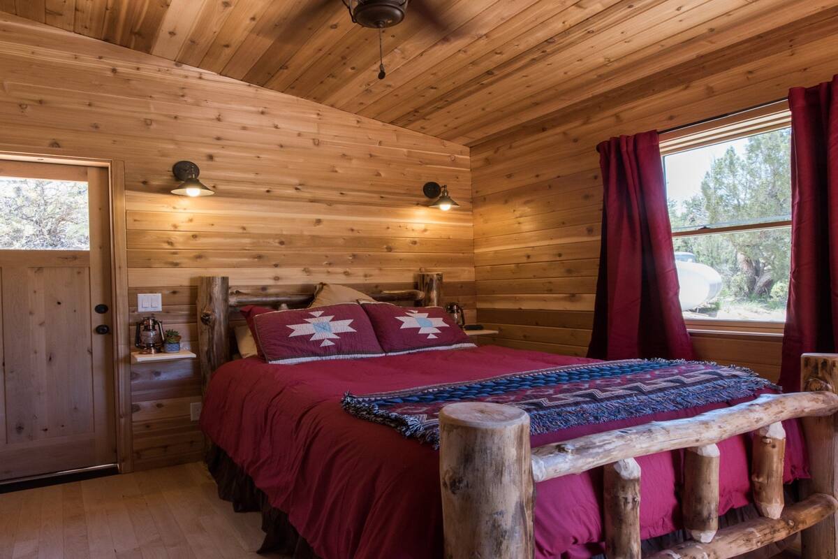 A cozy log cabin bedroom with a wooden bed, a window with red curtains, and a door with a small window.