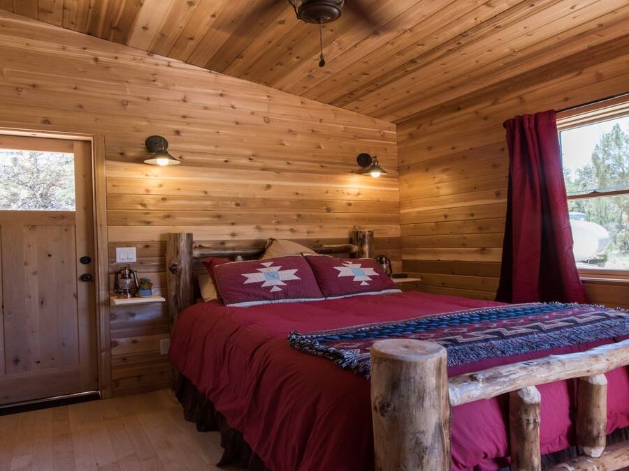 Een gezellige slaapkamer in een blokhut, met een houten bed, een raam met rode gordijnen en een deur met een klein raampje.