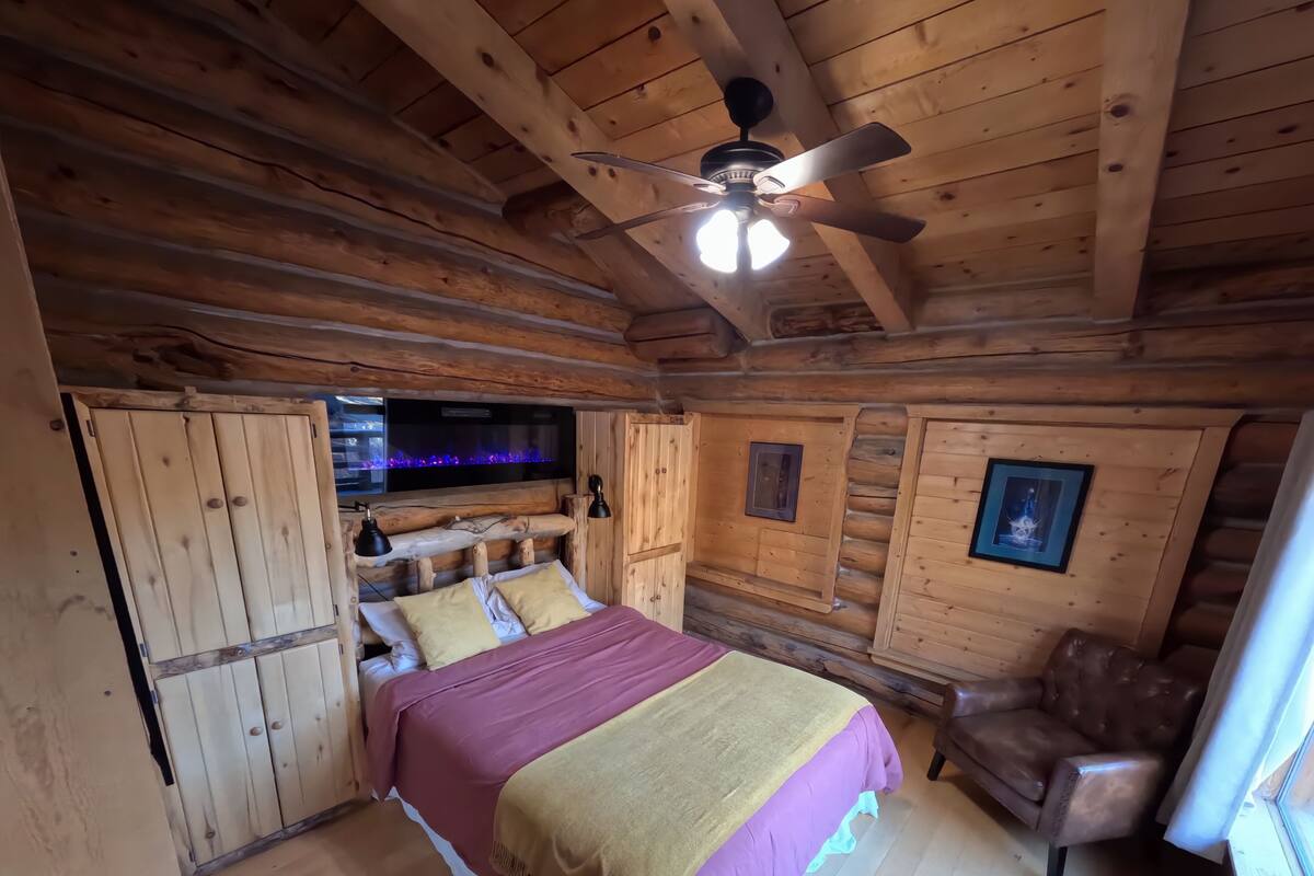 A log cabin bedroom with a bed, a ceiling fan, a mounted TV, and a leather chair.