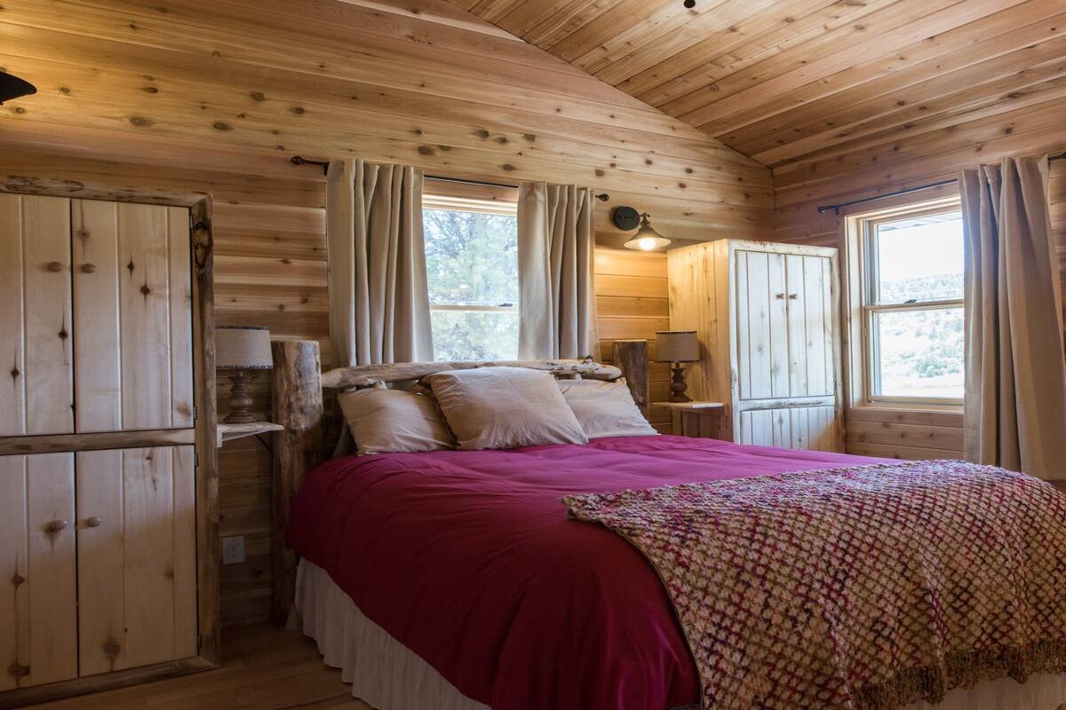 A wooden cabin bedroom with a bed, curtains, a nightstand, and a window.