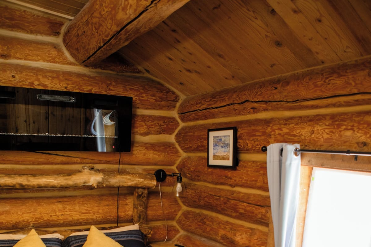 A log cabin bedroom with a bed, pillows, a mounted heater, a picture on the wall, and a window with curtains.