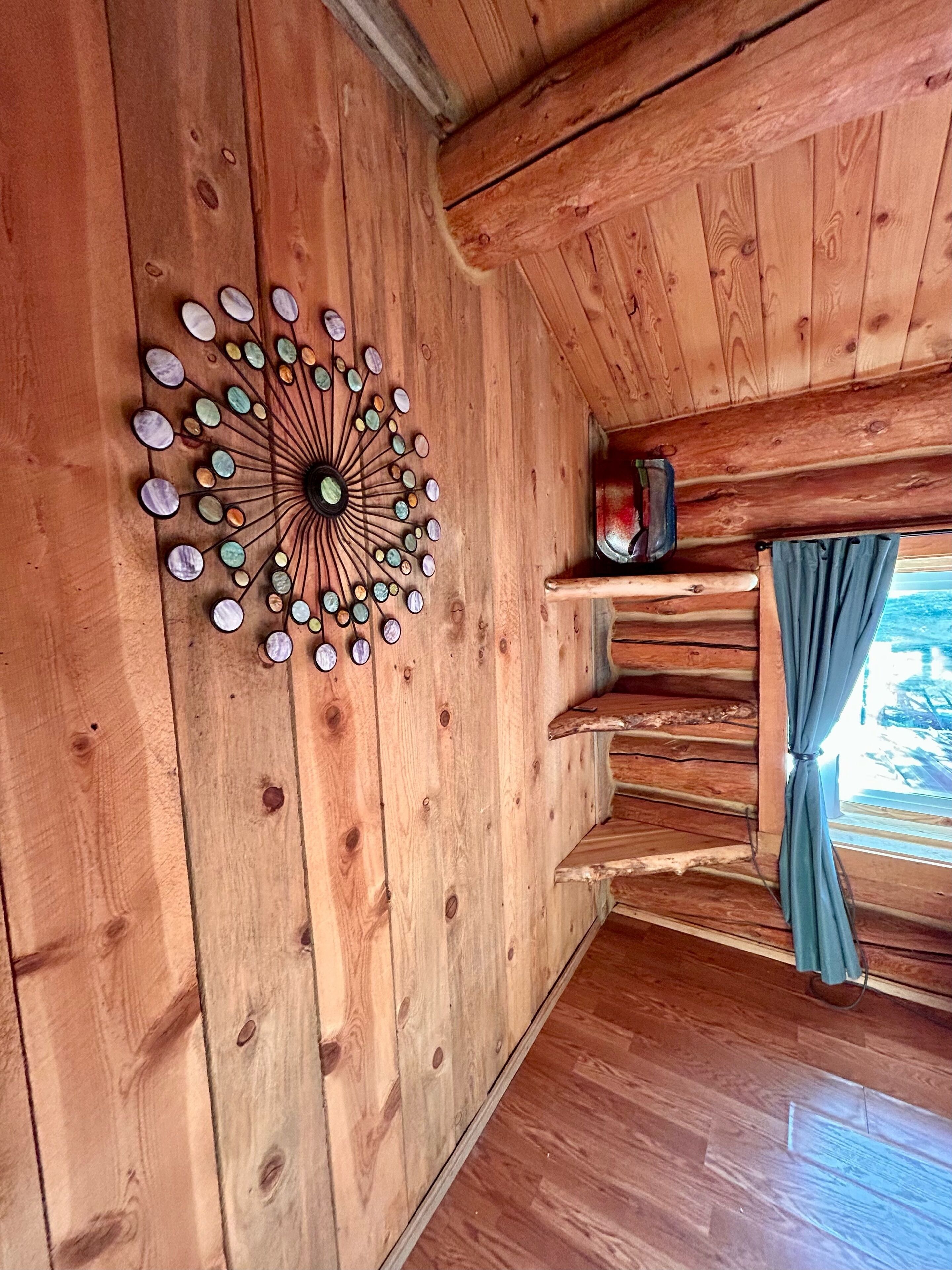 Ein Holzblockhaus-Interieur mit einer runden Wanddekoration aus Steinen, einem kleinen Regal mit einem roten Gegenstand und einem Fenster mit Vorhängen.