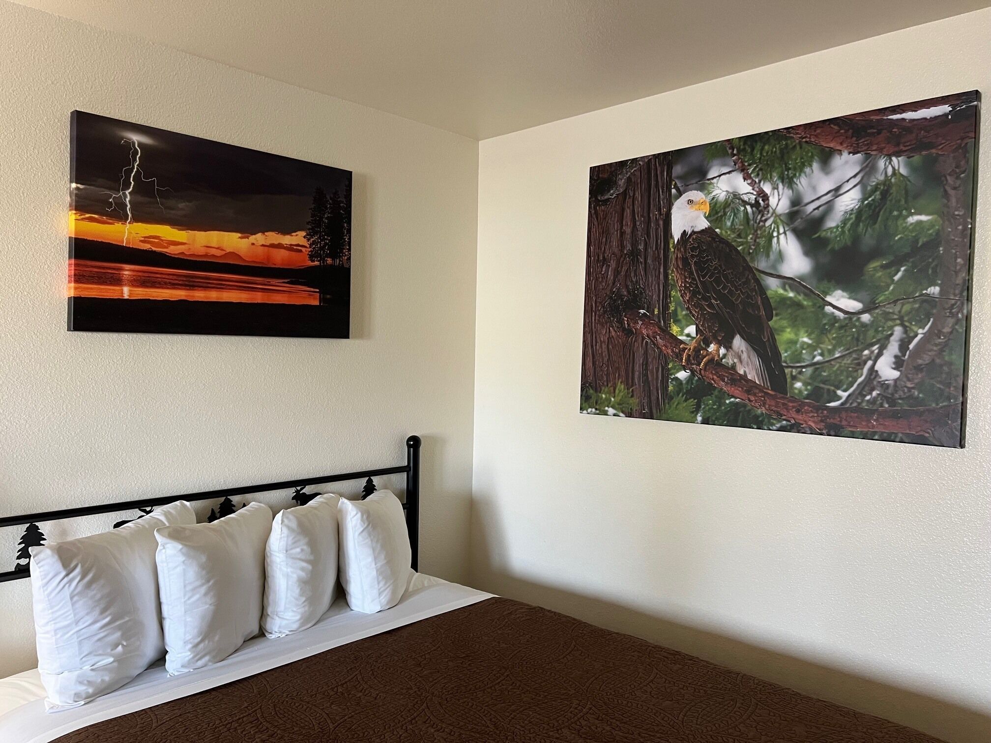 A bedroom with a bed, white pillows, and two wall-mounted paintings: one of a lightning storm and one of an eagle perched on a tree branch.