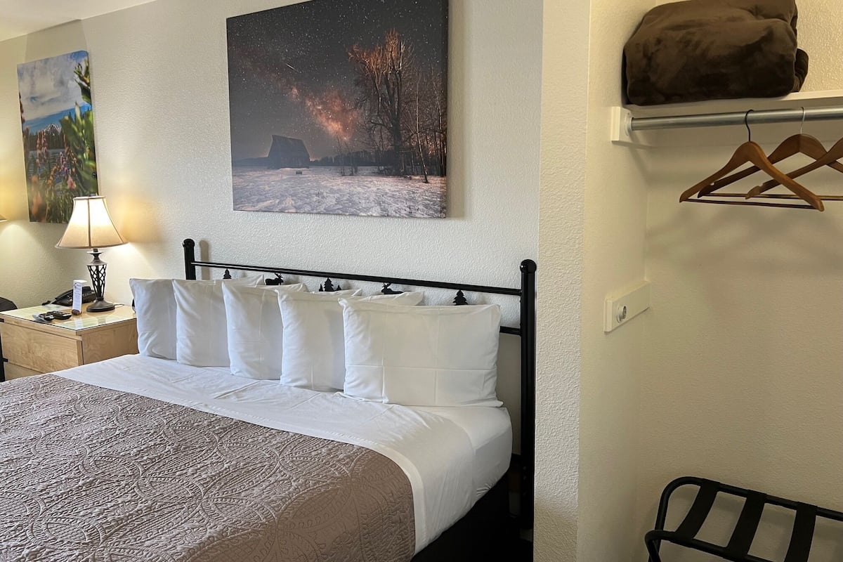 A neatly made bed with white linens and a patterned bedspread, a bedside lamp, a painting of a snowy landscape, and a coat hanging on a rack.