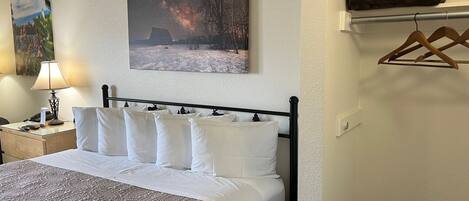 A neatly made bed with white linens and a patterned bedspread, a bedside lamp, a painting of a snowy landscape, and a coat hanging on a rack.