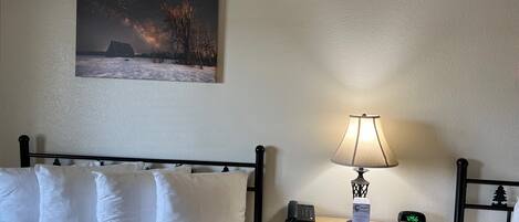 A hotel room with a bed, a nightstand with a lamp, a phone, a clock, and a painting of a snowy landscape.