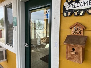 Un escaparate con una puerta verde, un cartel de 'Welcome', un buzón para 'Key Drop' y un adhesivo en la ventana que dice '¡Cute Little Thing In Chester!'.