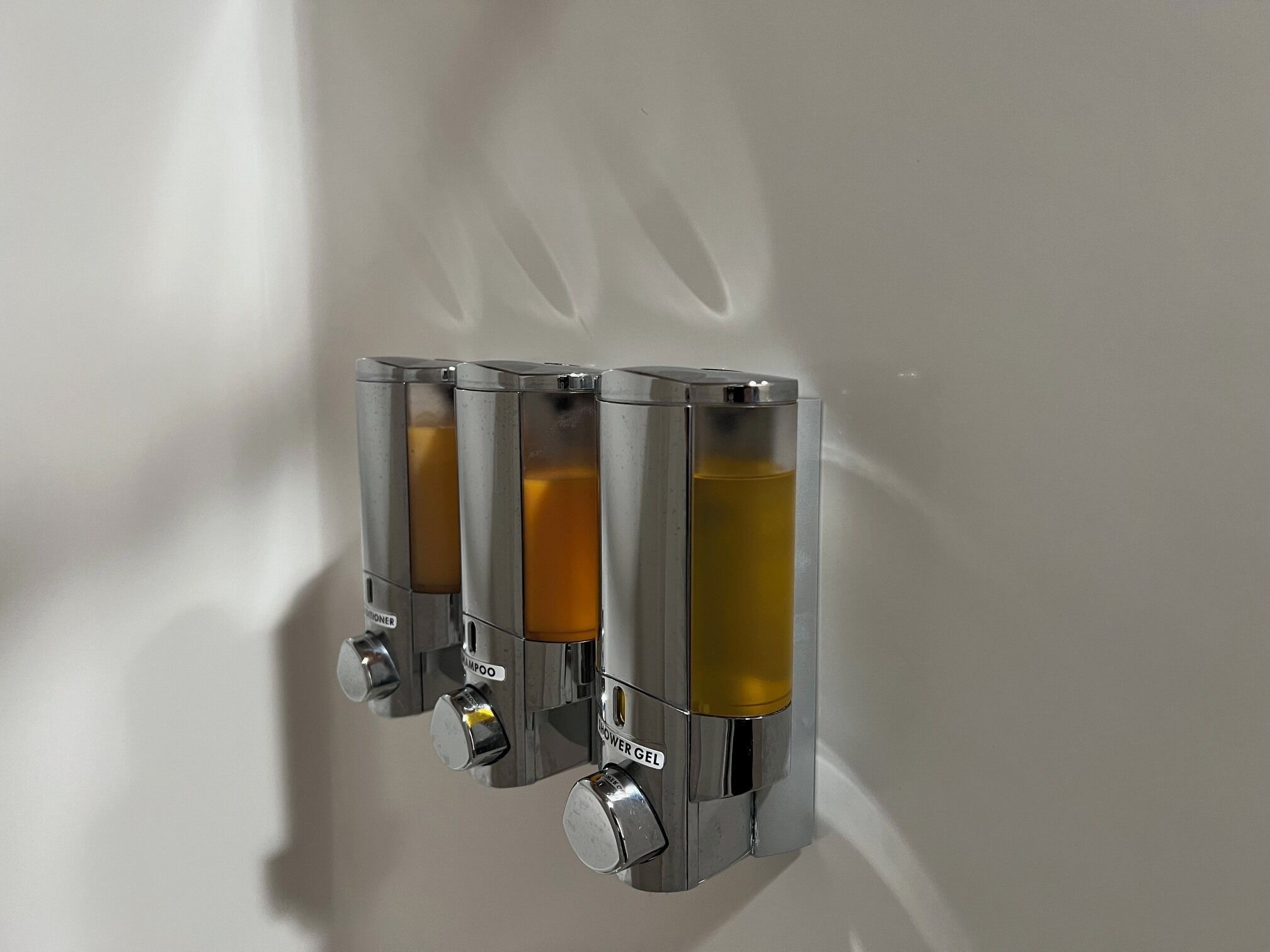 Three metallic dispensers with labels for shampoo and shower gel mounted on a white wall.