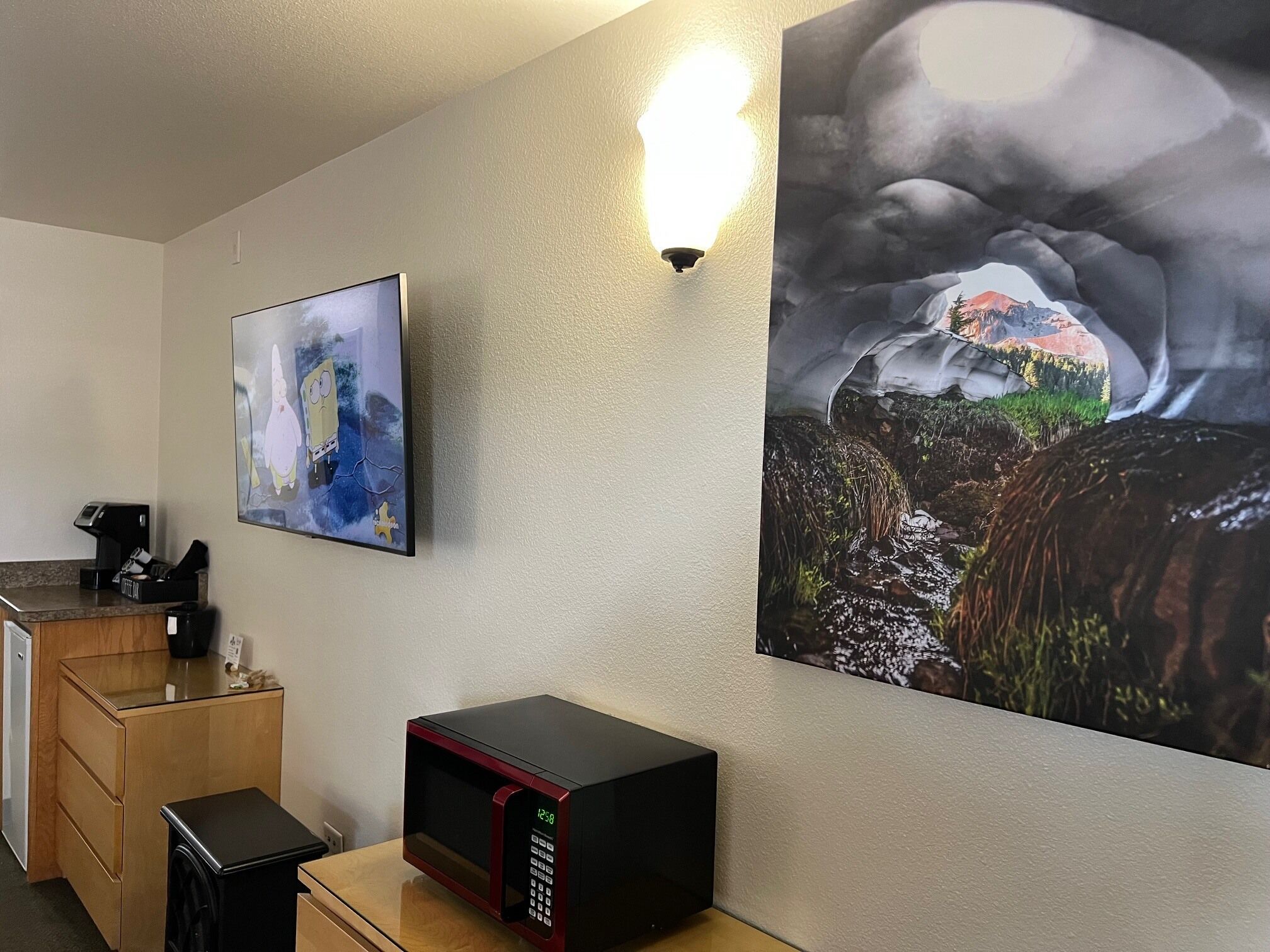 A hotel room with a microwave on a wooden table, a coffee machine, and a painting of a mountain landscape.