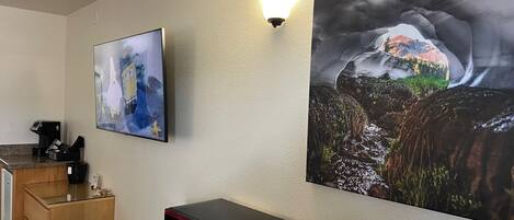 A hotel room with a microwave on a wooden table, a coffee machine, and a painting of a mountain landscape.