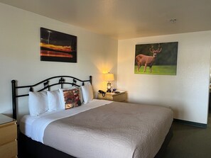 Una cama bien tendida con una colcha estampada, una mesita de noche con lámpara, y dos cuadros en la pared que representan un paisaje montañoso y un ciervo.