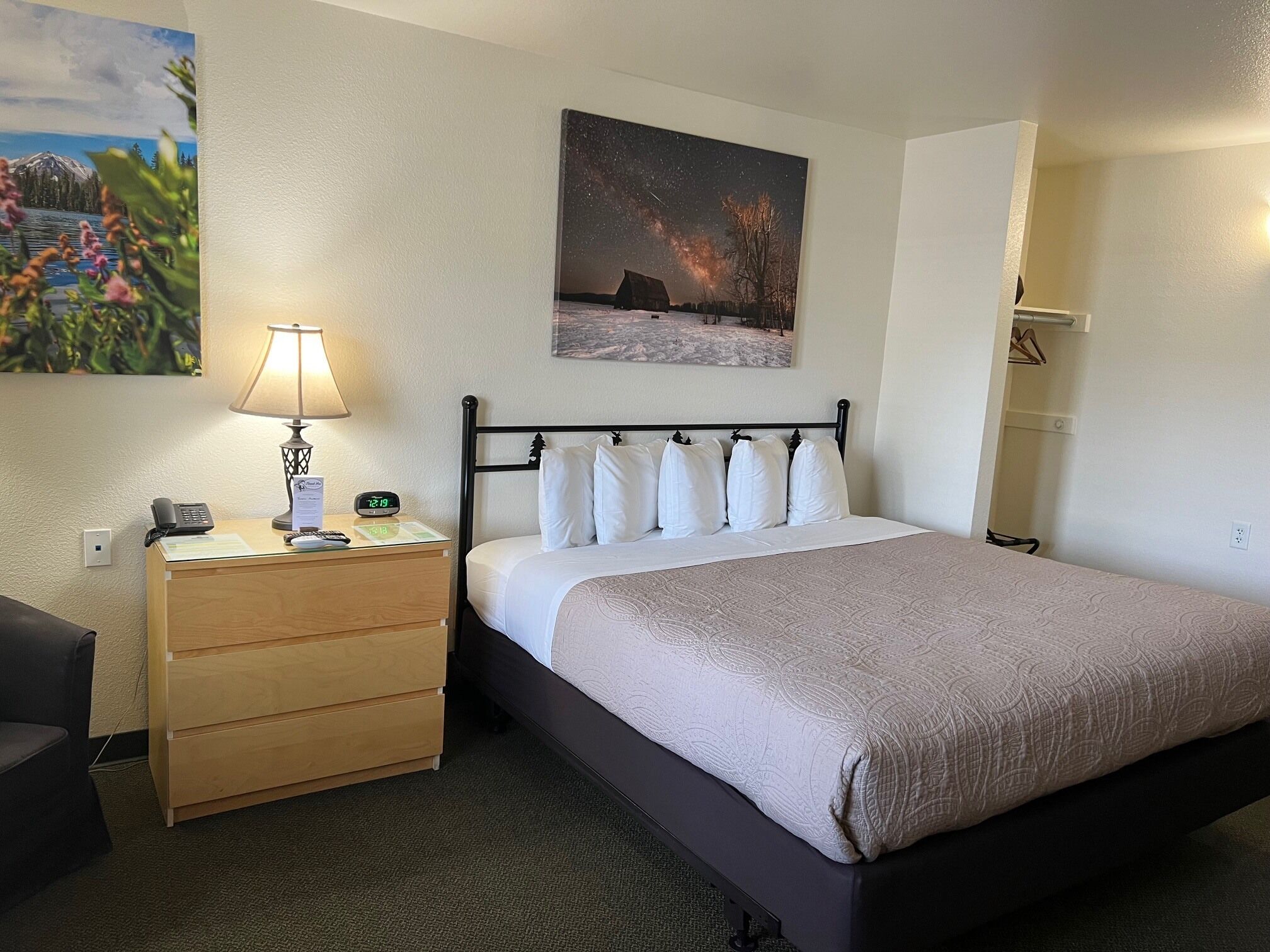 A neatly arranged hotel room with a bed, bedside table with a lamp, a clock, and a painting above the bed.