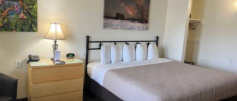 A neatly arranged hotel room with a bed, bedside table with a lamp, a clock, and a painting above the bed.