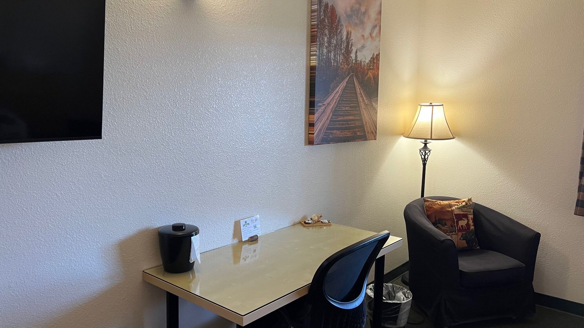 A small hotel room with a single table, a chair, a lamp, and a trash bin.