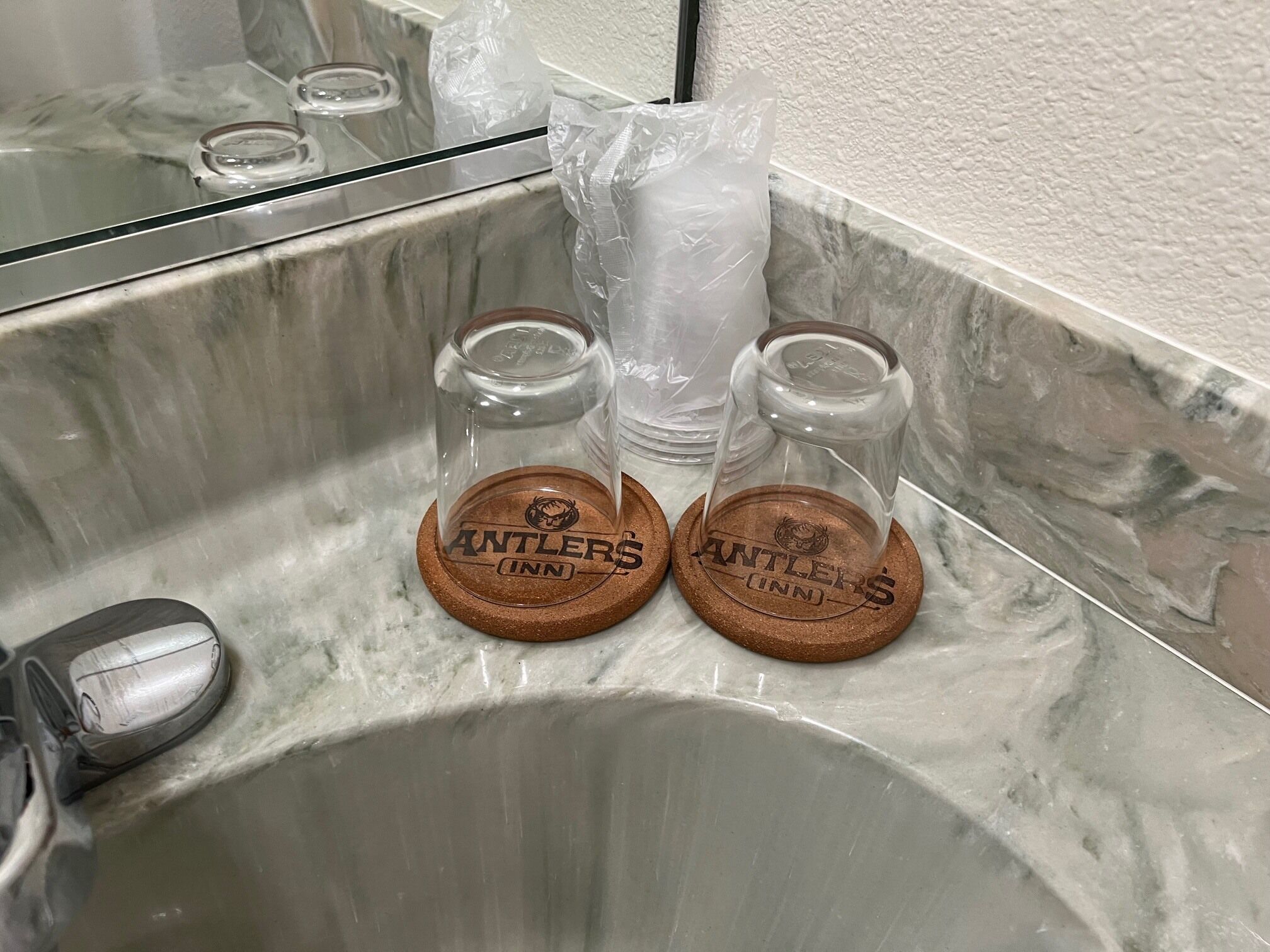 A bathroom sink with two coasters labeled "Antlers Inn" and a glass jar wrapped in plastic.