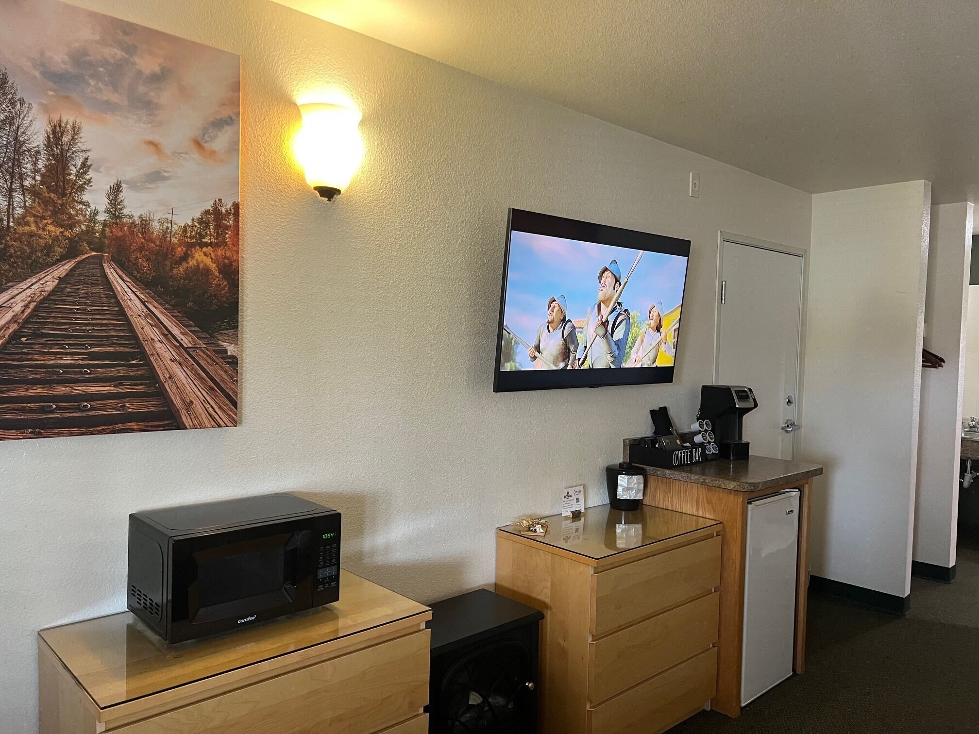A hotel room with a wooden desk, a microwave, a coffee maker, a TV, and a painting of a bridge.