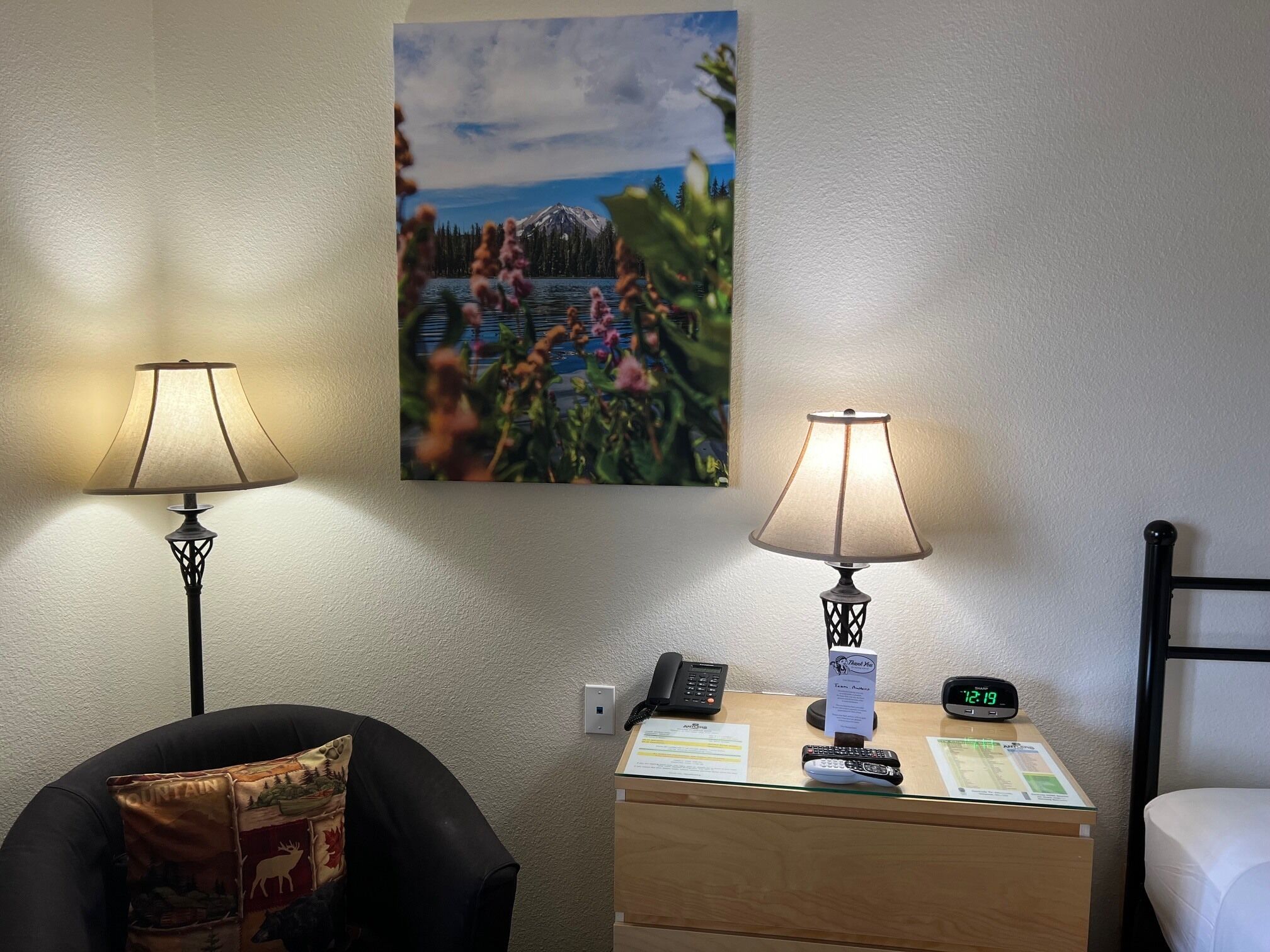 Habitación de hotel con dos lámparas, un cuadro de un paisaje montañoso, un escritorio con teléfono, un reloj y una cama.