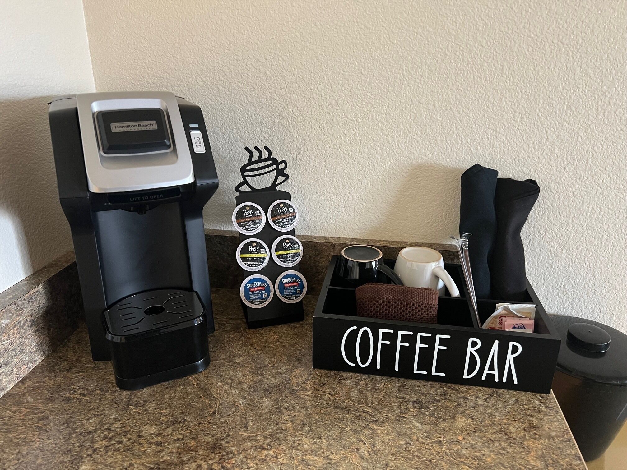 Una estación de café con una cafetera, un letrero de bar de café y un porta tazas con cápsulas de café.