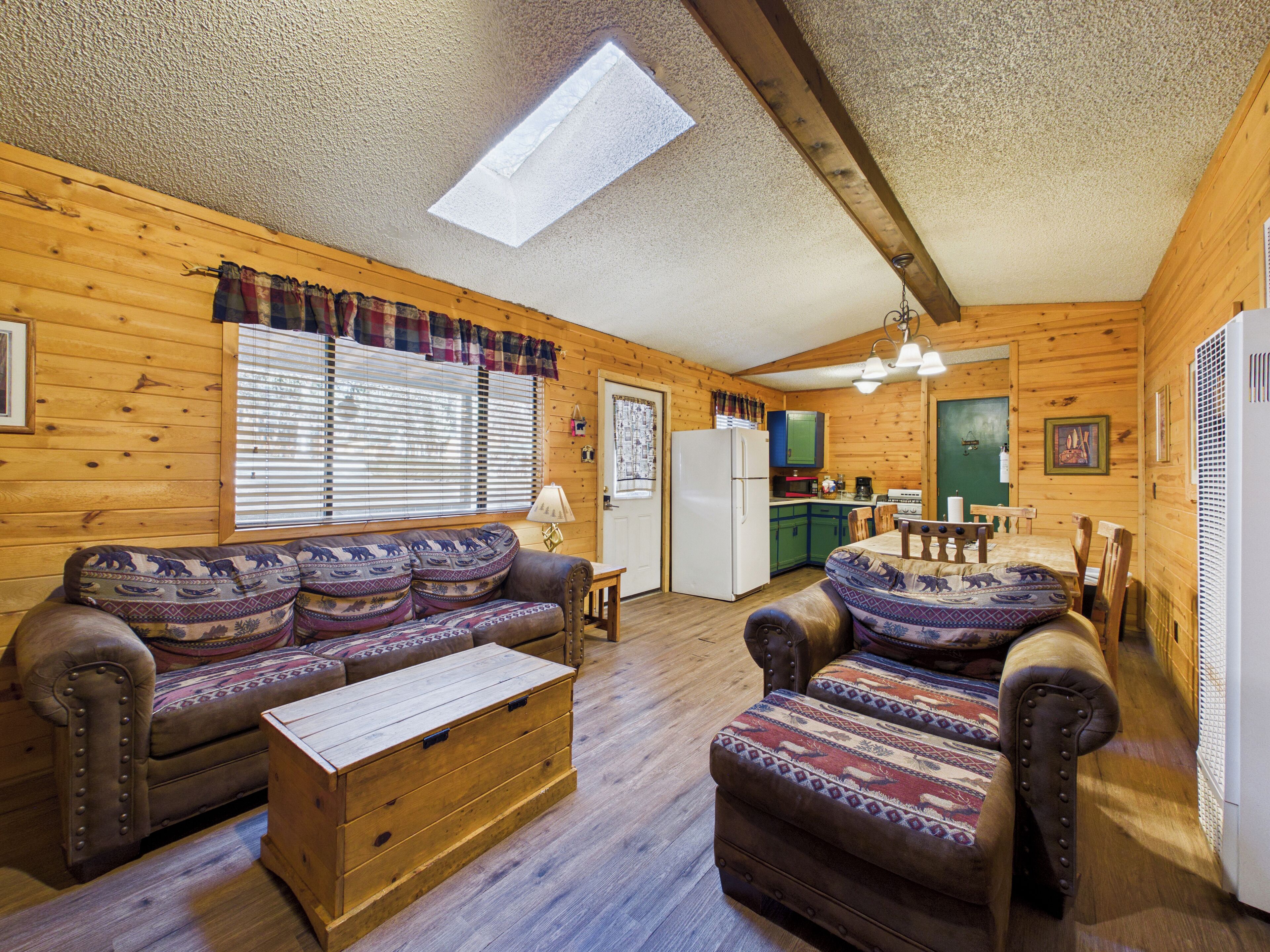 A cozy log cabin interior with a wooden sofa, a matching armchair, a wooden chest of drawers, a dining table with chairs, a kitchen area with a refrigerator, and a skylight.