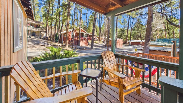 A wooden deck with Adirondack chairs and a table, overlooking a forested area with cabins. - Whispering Pine Cabins (Ruidoso)