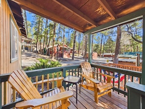 Una terraza de madera con sillas Adirondack y una mesa, con vistas a un área boscosa con cabañas.