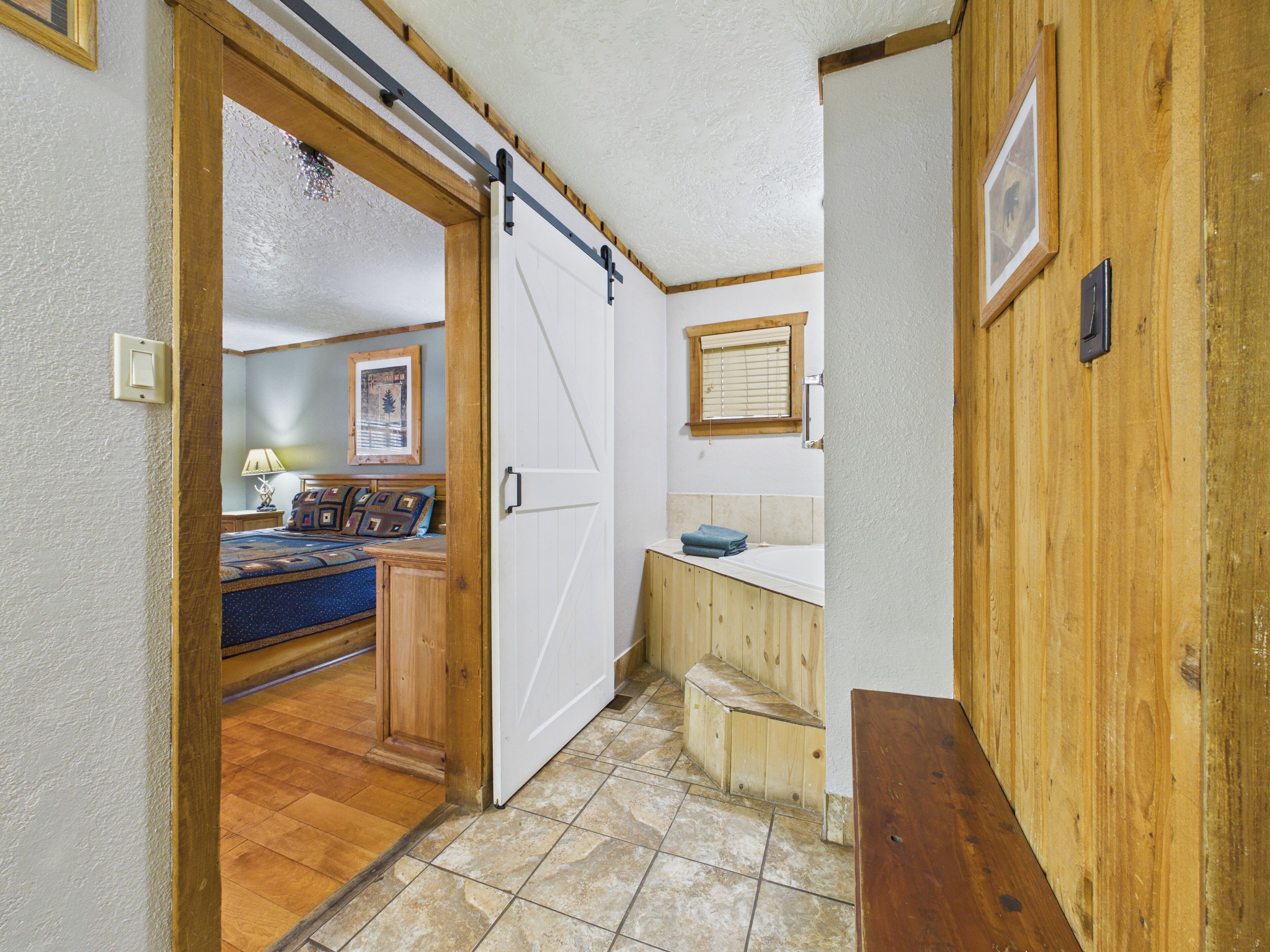 A narrow hallway with a wooden door leading to a bedroom with a bed and a nightstand.